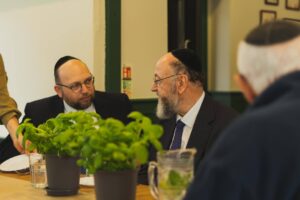 Chief Rabbi Mirvis sitting at the table with another person. There is a basil plant on the table and the two of them are looking at each other.