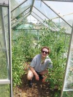Felix Wilk, a white person with short brown hair and sunglasses, crouching in a greenhouse smiling